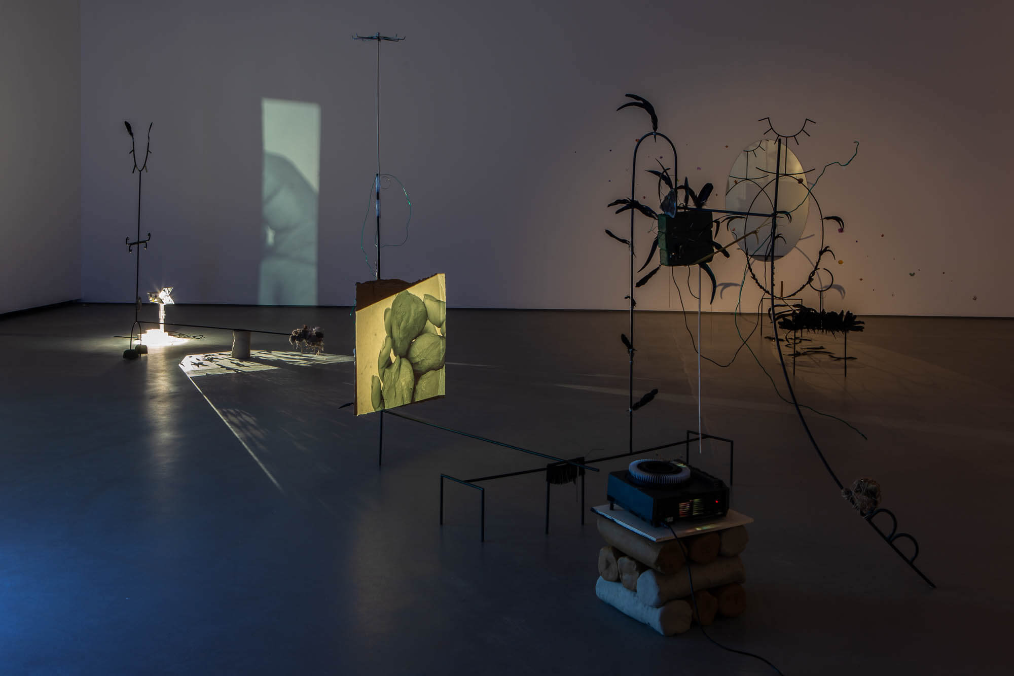 An installation in the gallery of many components, including metal rods with feather-like appendages