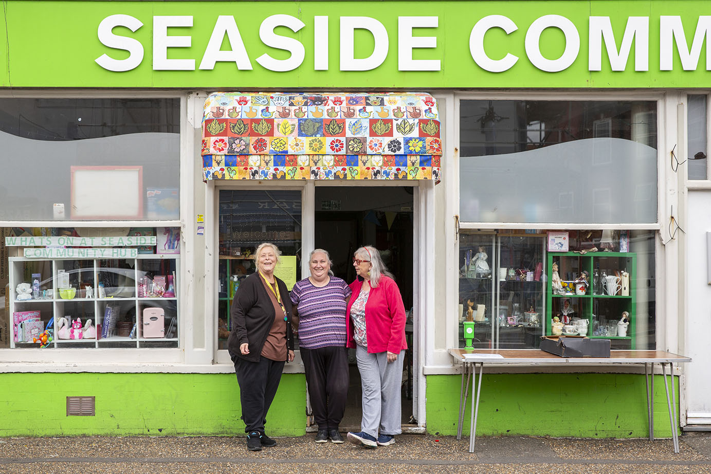 Three women standing outside Seaside Community Centre, which has an awning made from colourful fabric. The fabric design is made up of tiles with symbols including palm trees, moons, sun and ice cream. 