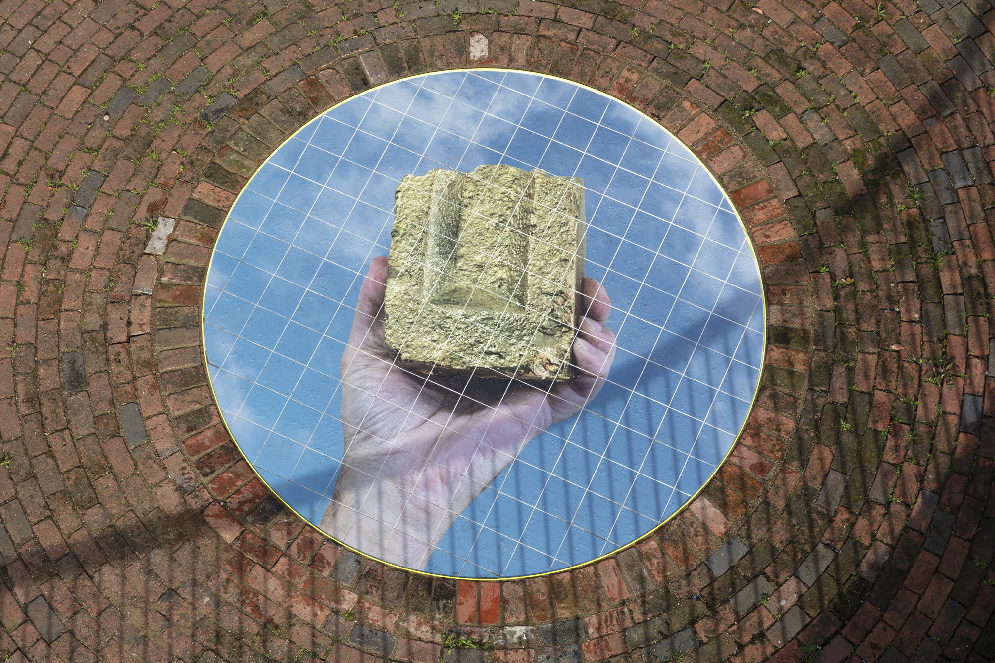 A photo of a hand holding a bronze object against a cloudy sky, printed on tiles which are embedded in the middle of a round brick formation in the ground. 